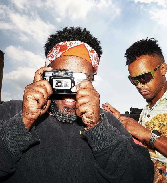 Two adult men are outdoors during the daytime; one is taking a photo using a vintage camera while the other poses in the background.