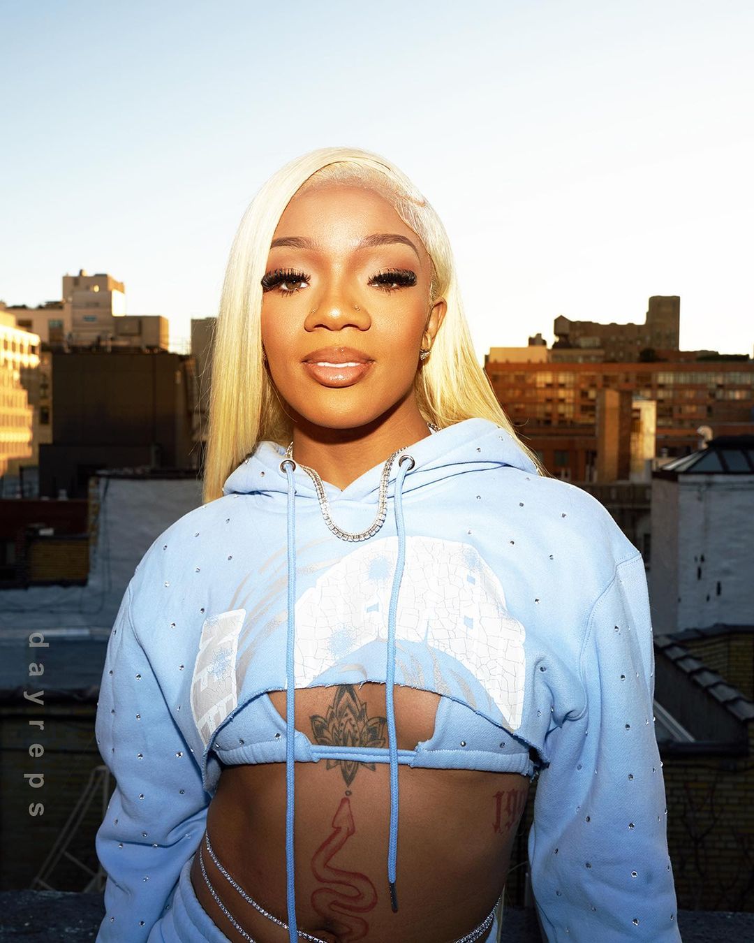 Portrait of a blonde woman wearing a blue cropped hoodie and a chain belt, standing on a rooftop with cityscape in the distance.