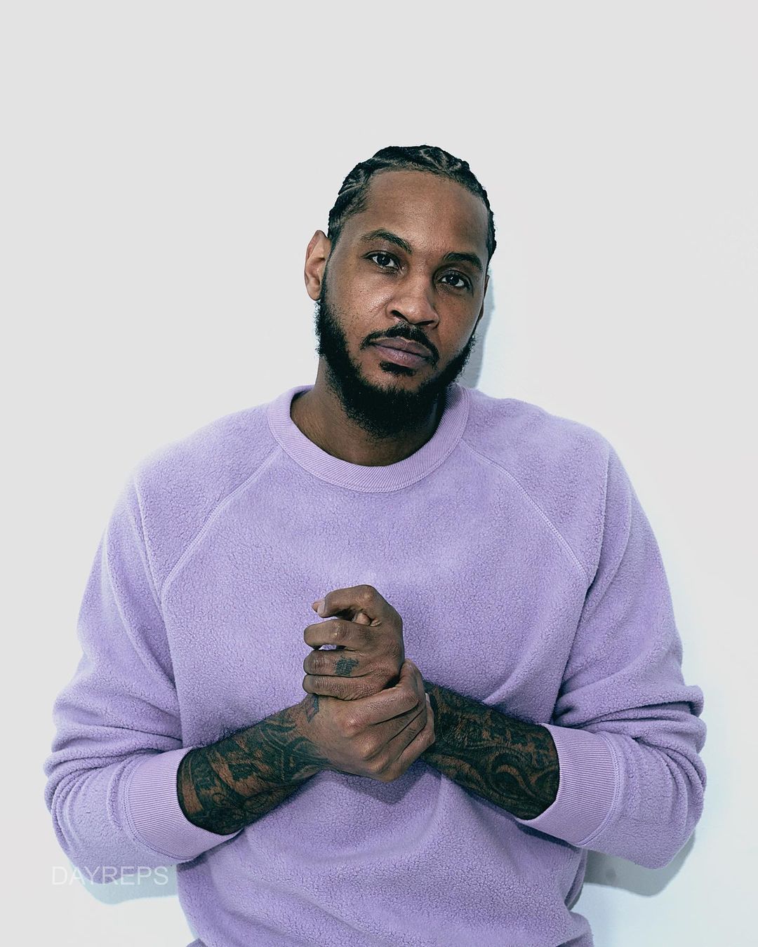 Portrait of a Black adult male wearing a textured light purple sweater standing indoors against a white wall; he has cornrows and a beard.