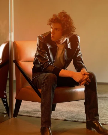 Young man with curly hair wearing a leather jacket seated in a brown armchair in Los Angeles by photographer Kareem Black