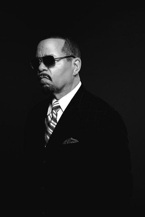 Monochrome portrait of Ice T in a dark suit, striped tie, and sunglasses against a black background.