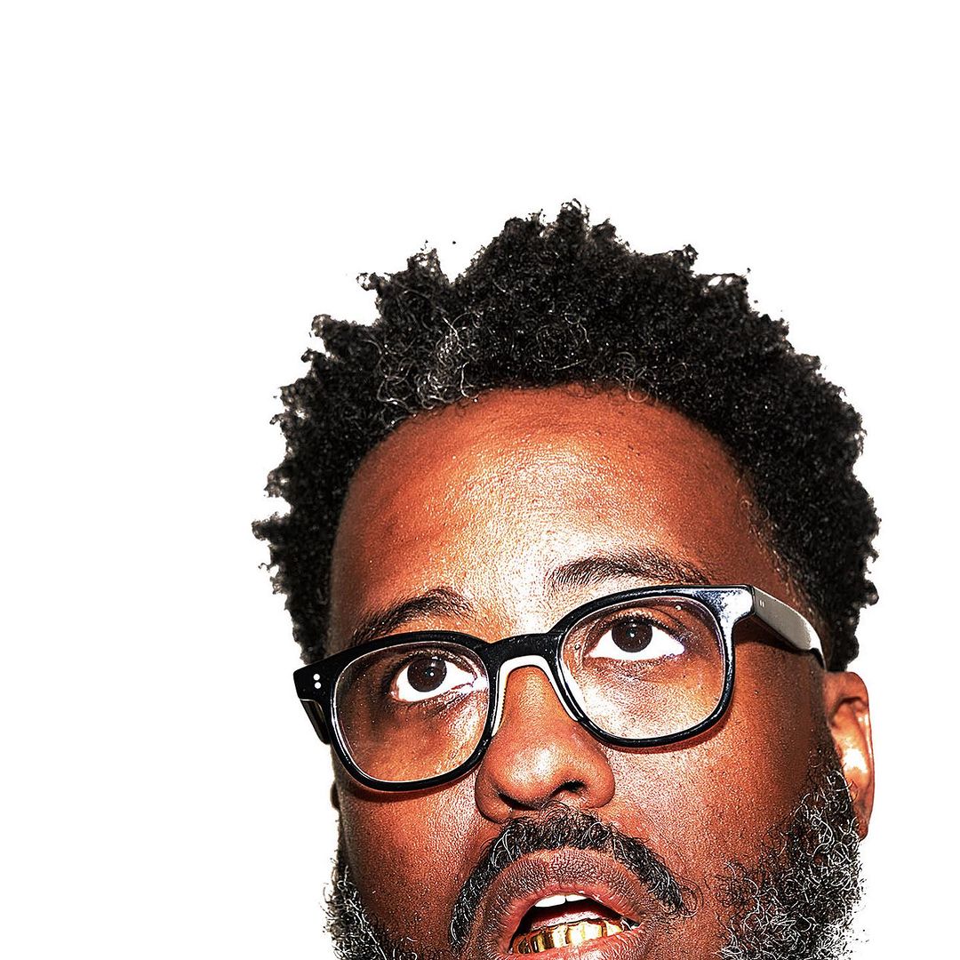 Close-up portrait of an adult man with dark skin, glasses, and a beard, looking upwards against a bright white background.