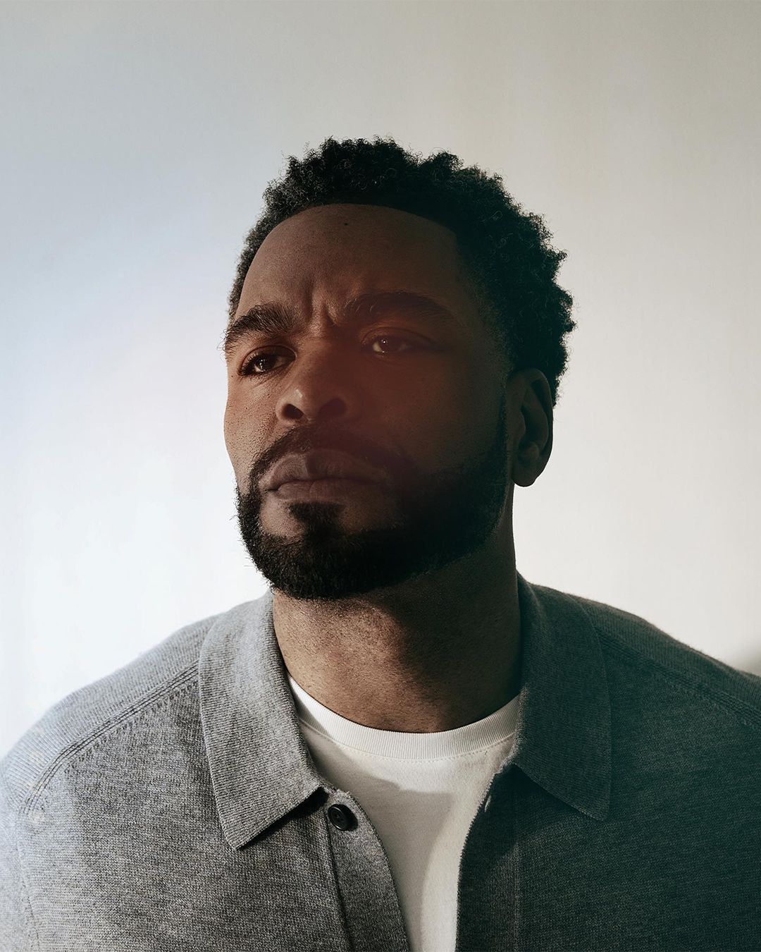Portrait of an adult Black man with a beard wearing a gray shirt and white undershirt against a bright background looking slightly up.