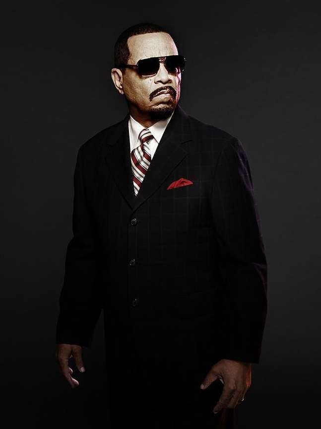 Portrait of Ice-T in a dark suit, tie, and sunglasses against a dark gray backdrop, showcasing a formal and serious demeanor.