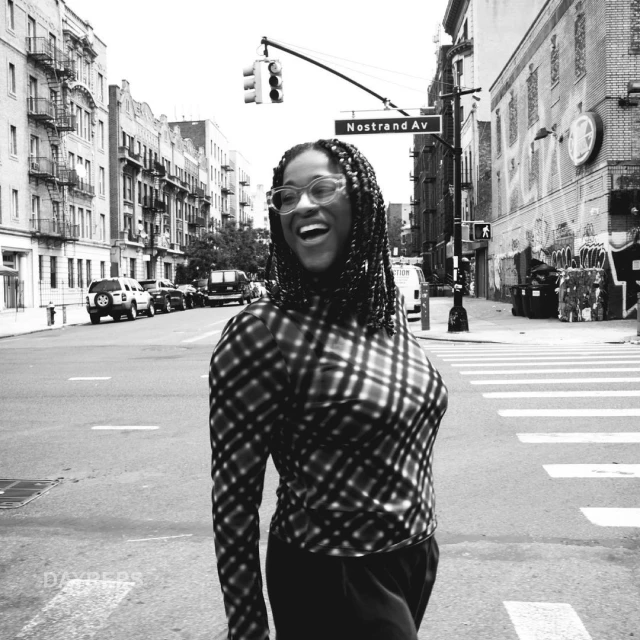 A smiling African-American adult female with braided hair is wearing glasses and a plaid top on an urban street with buildings and traffic.
