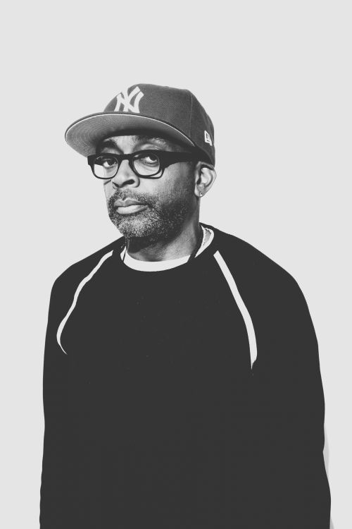 Portrait of Spike Lee wearing a baseball cap and thick-framed glasses in a black and white monochrome studio shot