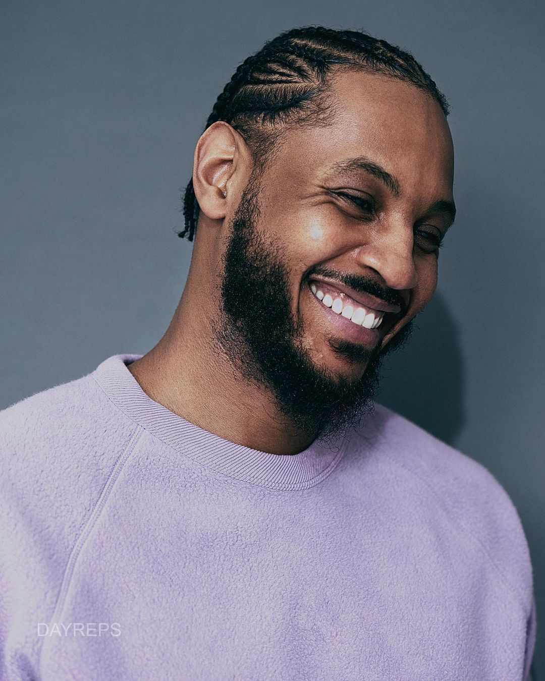 Close-up portrait of an adult male with braided hair smiling and wearing a light purple sweater against a grey backdrop.