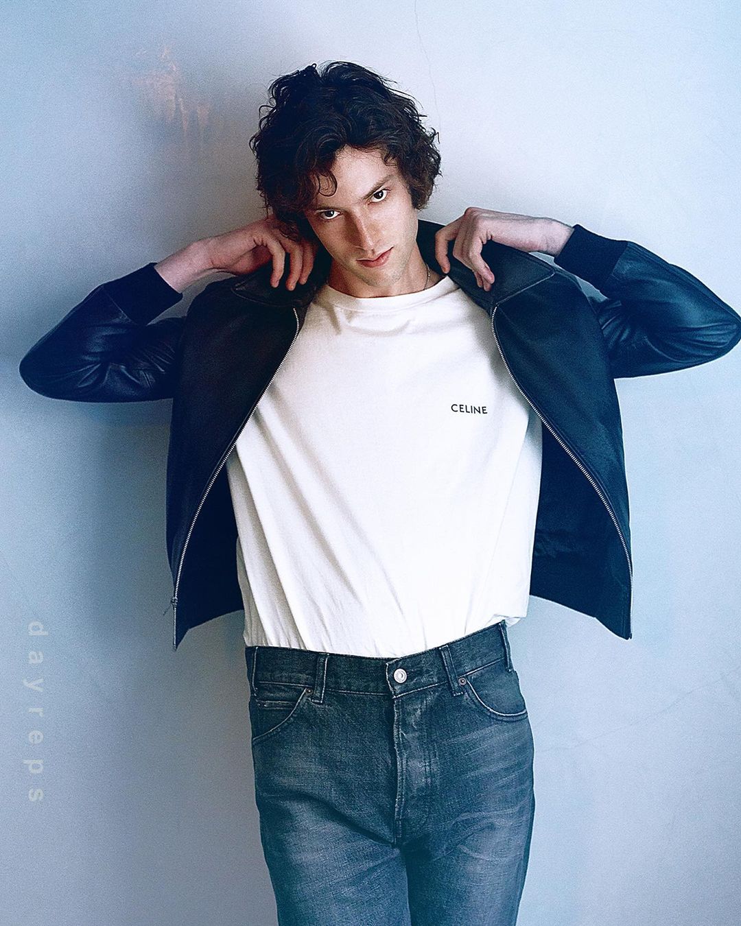 Portrait of a man with dark curly hair wearing a white t-shirt, dark bomber jacket, and denim jeans against a grey wall.