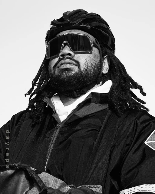 Monochrome portrait of a bearded adult male wearing a helmet and sunglasses, looking up with a neutral expression against a white background.