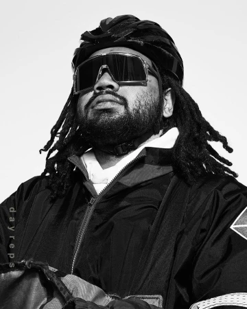 Monochrome portrait of a bearded adult male wearing a helmet and sunglasses, looking up with a neutral expression against a white background.