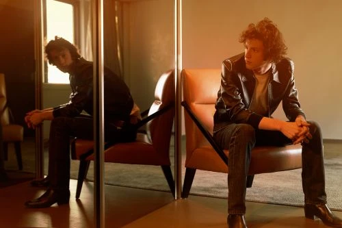 A man with curly hair wearing a leather jacket and dark jeans is seated in an armchair, reflected in a mirror.