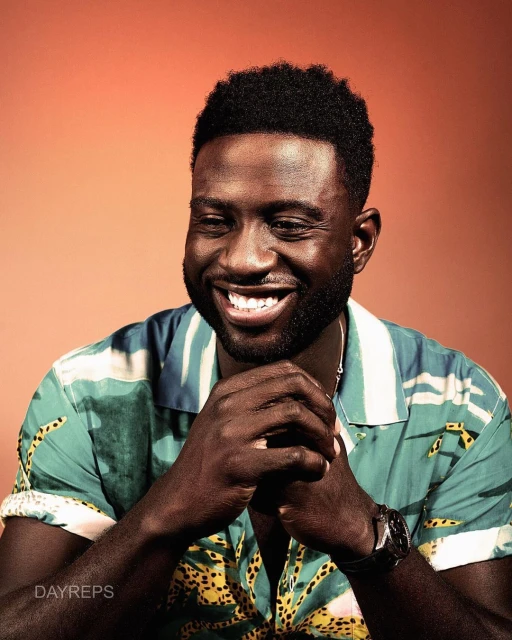 Smiling man with dark skin and short hair in a patterned shirt against an orange background in a portrait shot with his hands clasped.