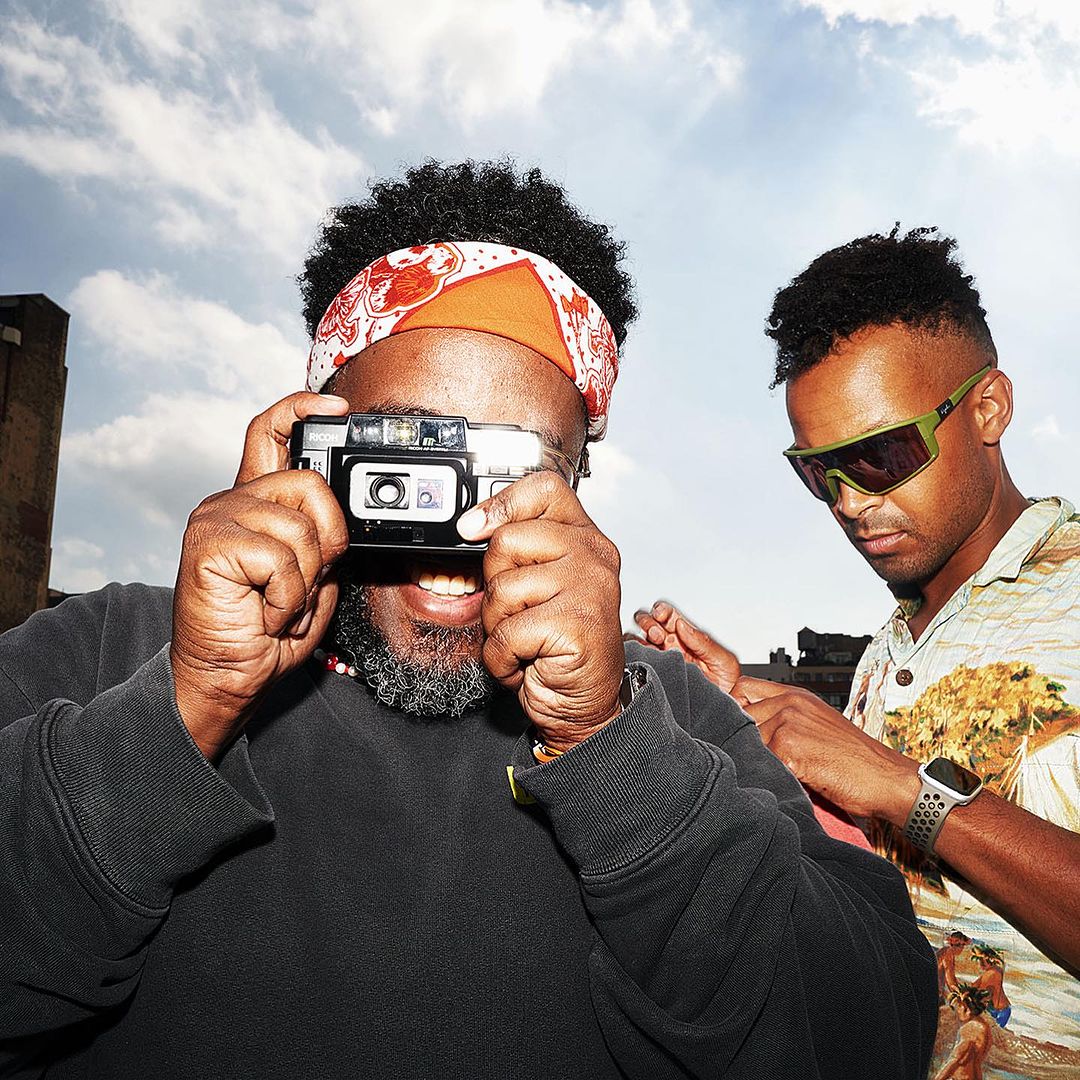 Two black men outdoors with a bright sky background, one holding a compact camera up to his eye while smiling, wearing a black sweatshirt and a patterned red and white headband.
