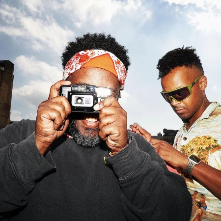 Two black men outdoors with a bright sky background, one holding a compact camera up to his eye while smiling, wearing a black sweatshirt and a patterned red and white headband.