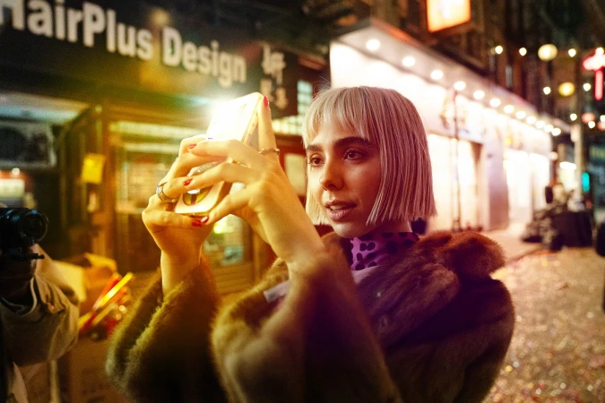 A young adult woman takes a photo with her smartphone in a city street setting at night, wearing a fur coat. Storefronts illuminated.