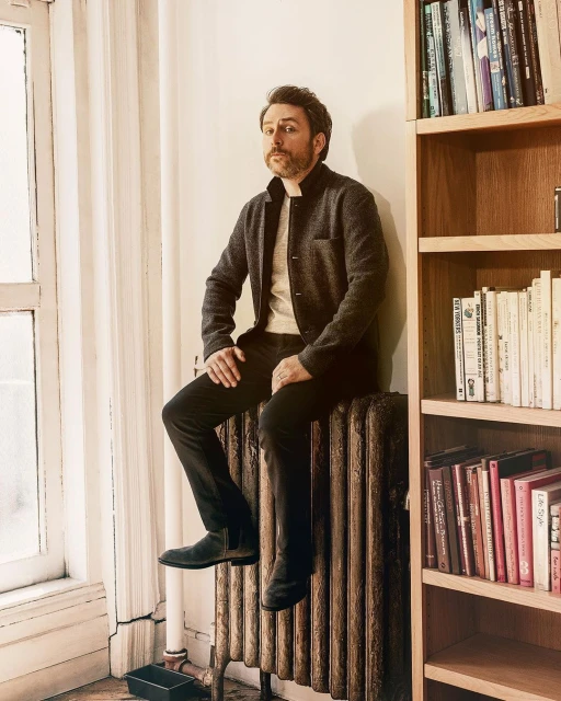 Adult man with beard sitting on radiator in front of window and bookshelf in casual gray jacket, cream shirt, dark pants and black boots.
