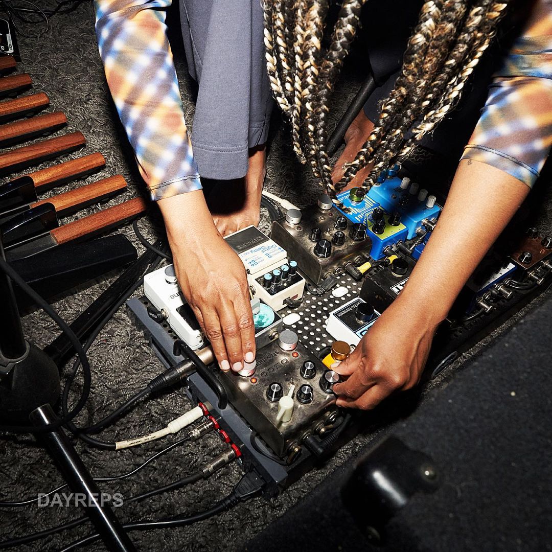 Musician adjusts a pedalboard with various effects pedals, cables, and a xylophone-like instrument partially visible in the background.