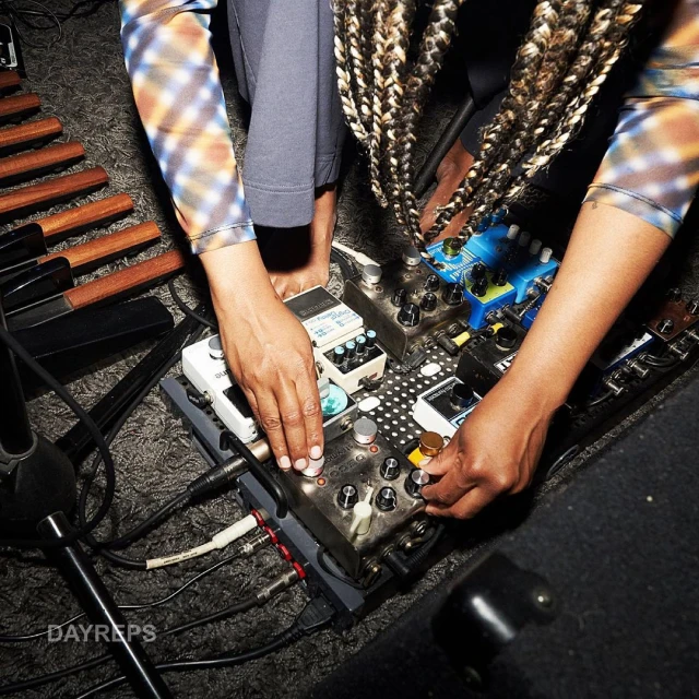 Musician adjusts a pedalboard with various effects pedals, cables, and a xylophone-like instrument partially visible in the background.