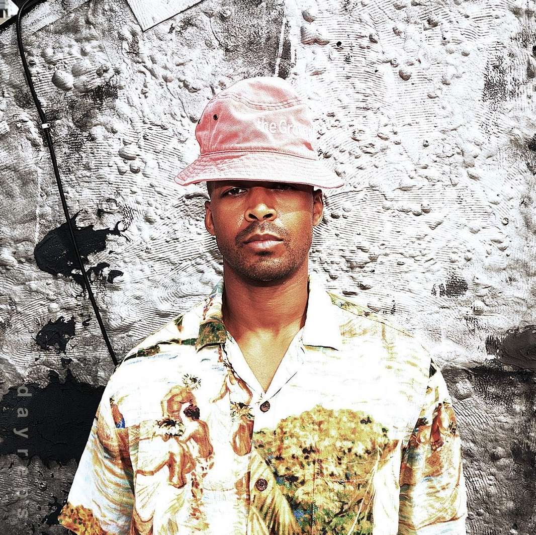 Close-up portrait of an adult male wearing a hat and patterned short-sleeve shirt, against a textured gray and black backdrop.