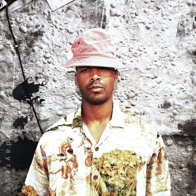 Close-up portrait of an adult male wearing a hat and patterned short-sleeve shirt, against a textured gray and black backdrop.