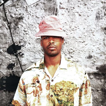 Close-up portrait of an adult male wearing a hat and patterned short-sleeve shirt, against a textured gray and black backdrop.