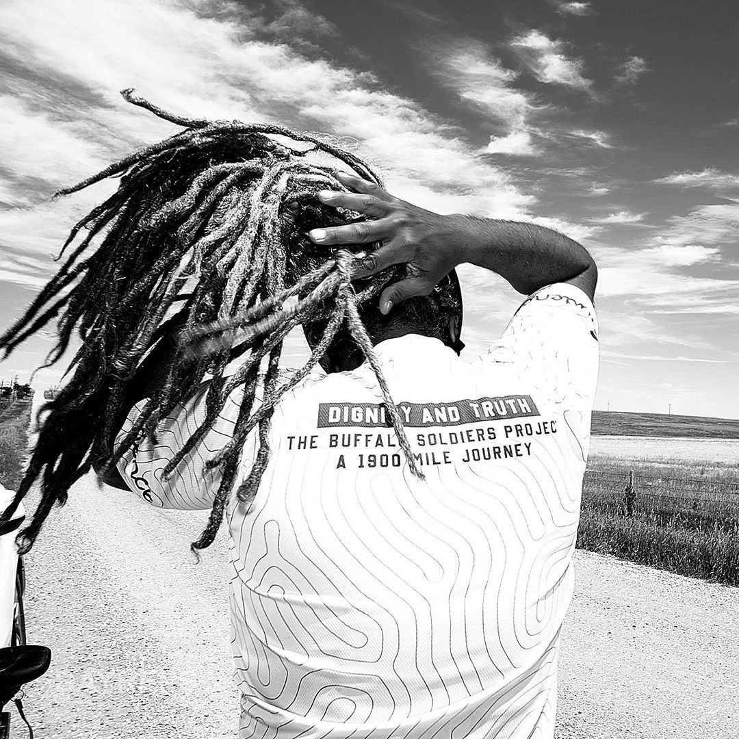 Black and white shot from behind of a person with dreadlocks wearing a topographic-patterned shirt in a rural, grassy setting.