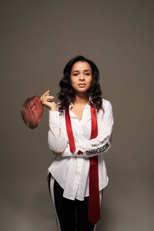 Adult female posing in a studio with a red tie, white shirt, and black and white striped pants, holding a brown hat.