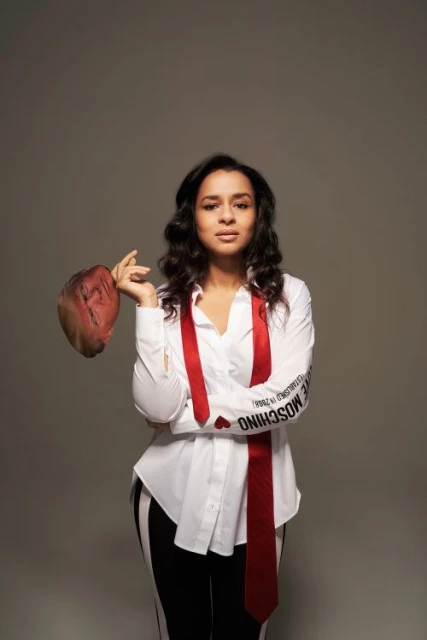Adult female posing in a studio with a red tie, white shirt, and black and white striped pants, holding a brown hat.