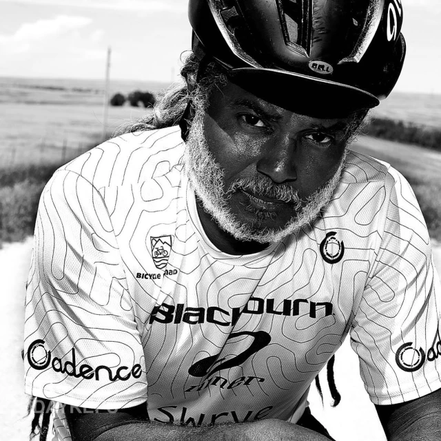 Close-up black and white portrait of a cyclist wearing a helmet and jersey with logos, posing outside on a rural road.