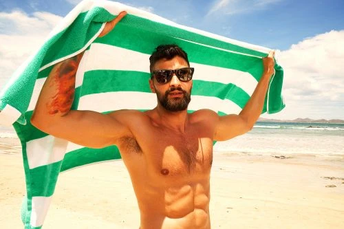 Shirtless man with sunglasses holding a green and white striped towel on a sandy beach