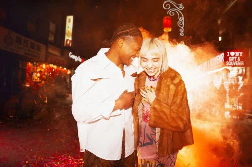 Stylish couple laughing and posing in a city street with a souvenir shop in the background. Nighttime scene, warm lighting.