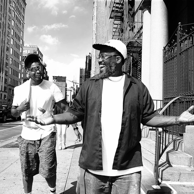 Two men walk on a city sidewalk in a black-and-white photograph. One man is laughing, wearing a hat and glasses. Buildings line the street.