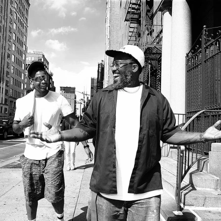 Two men walk on a city sidewalk in a black-and-white photograph. One man is laughing, wearing a hat and glasses. Buildings line the street.