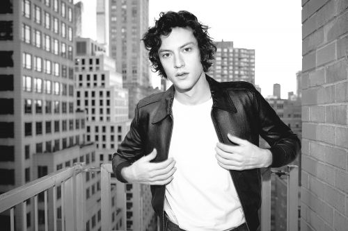 Monochrome portrait of a young adult man in a leather jacket standing on a balcony in a city with tall buildings in the background.