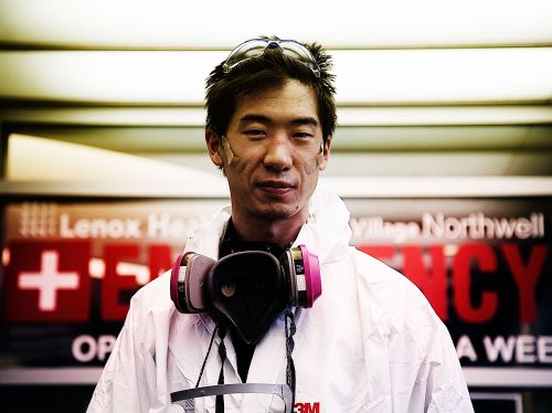 Asian adult wearing a white hazmat suit and a respirator, with safety goggles perched on their head, in an indoor setting.