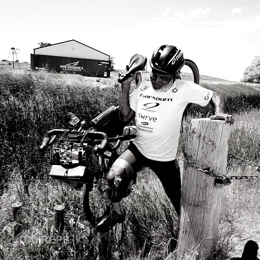 Athlete pauses with adaptive bike on rural road in black and white, wearing helmet and cycling jersey in outdoor setting.
