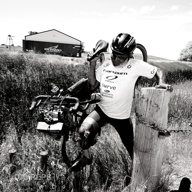 Athlete pauses with adaptive bike on rural road in black and white, wearing helmet and cycling jersey in outdoor setting.