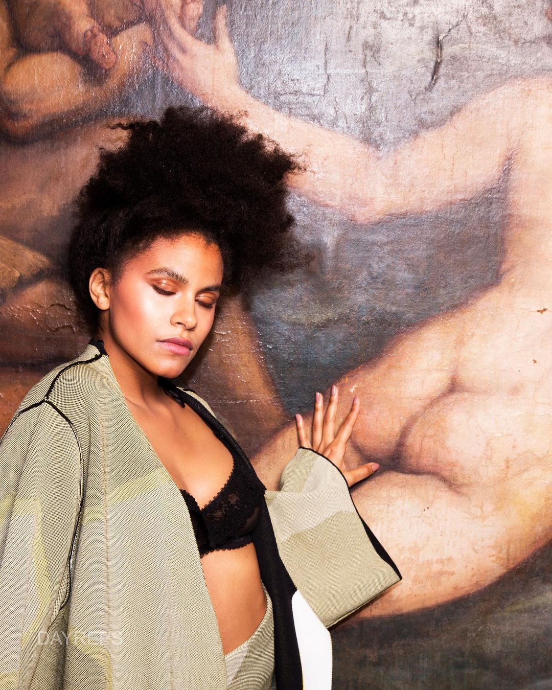 Adult woman with afro poses in front of an art background. She is wearing a jacket over a bra. Indoor, soft lighting, medium shot.