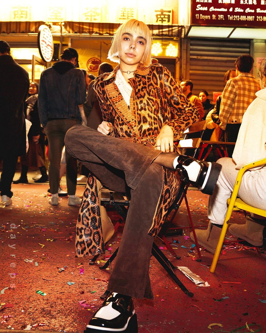Blonde woman in patterned coat sits in an outdoor, urban setting amidst confetti, wearing platform shoes and dark tights.