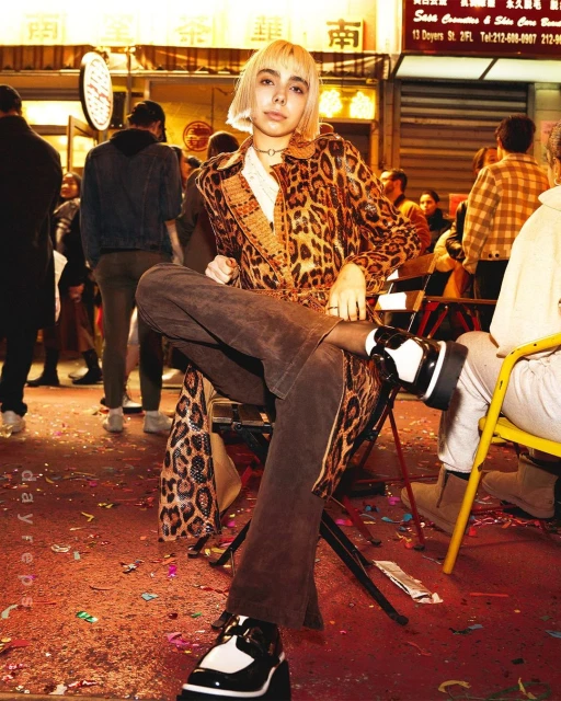 Blonde woman in patterned coat sits in an outdoor, urban setting amidst confetti, wearing platform shoes and dark tights.
