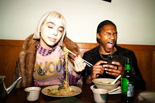 Two people, an adult female and an adult male, sit at a table with bowls and plates of noodles and a bottle of beer at an indoor restaurant.