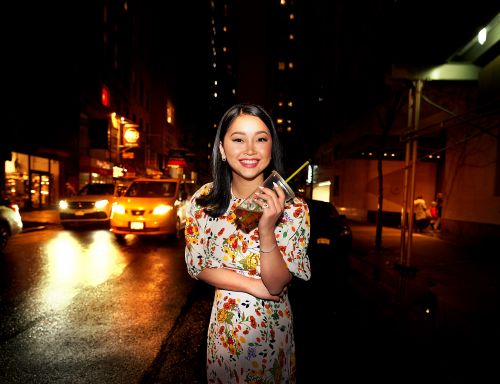 Woman smiles, holding drink, floral dress, night street scene, bright taxi, and cityscape lights create a vibrant urban setting.