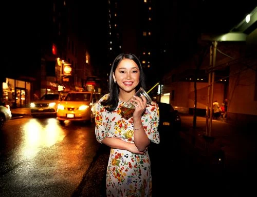 Woman smiles, holding drink, floral dress, night street scene, bright taxi, and cityscape lights create a vibrant urban setting.