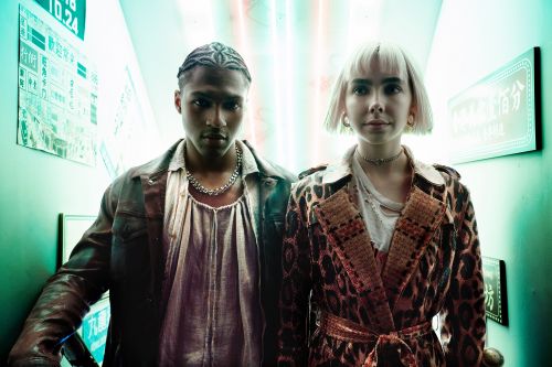 Close-up shot of a stylish couple in a brightly lit corridor. The male wears a leather jacket; the female, a leopard-print coat.