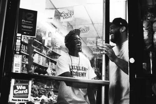 Black-and-white photo of two Black adult males laughing together in front of a store, captured with natural light and an eye-level angle.