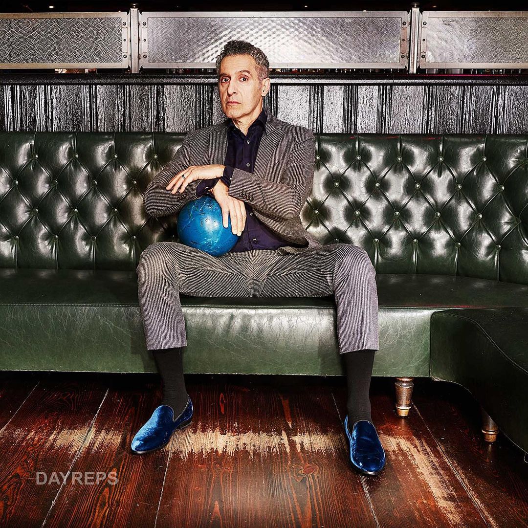 Adult male seated on a green tufted couch holding a blue bowling ball, wearing a gray suit and blue velvet loafers in an indoor setting.