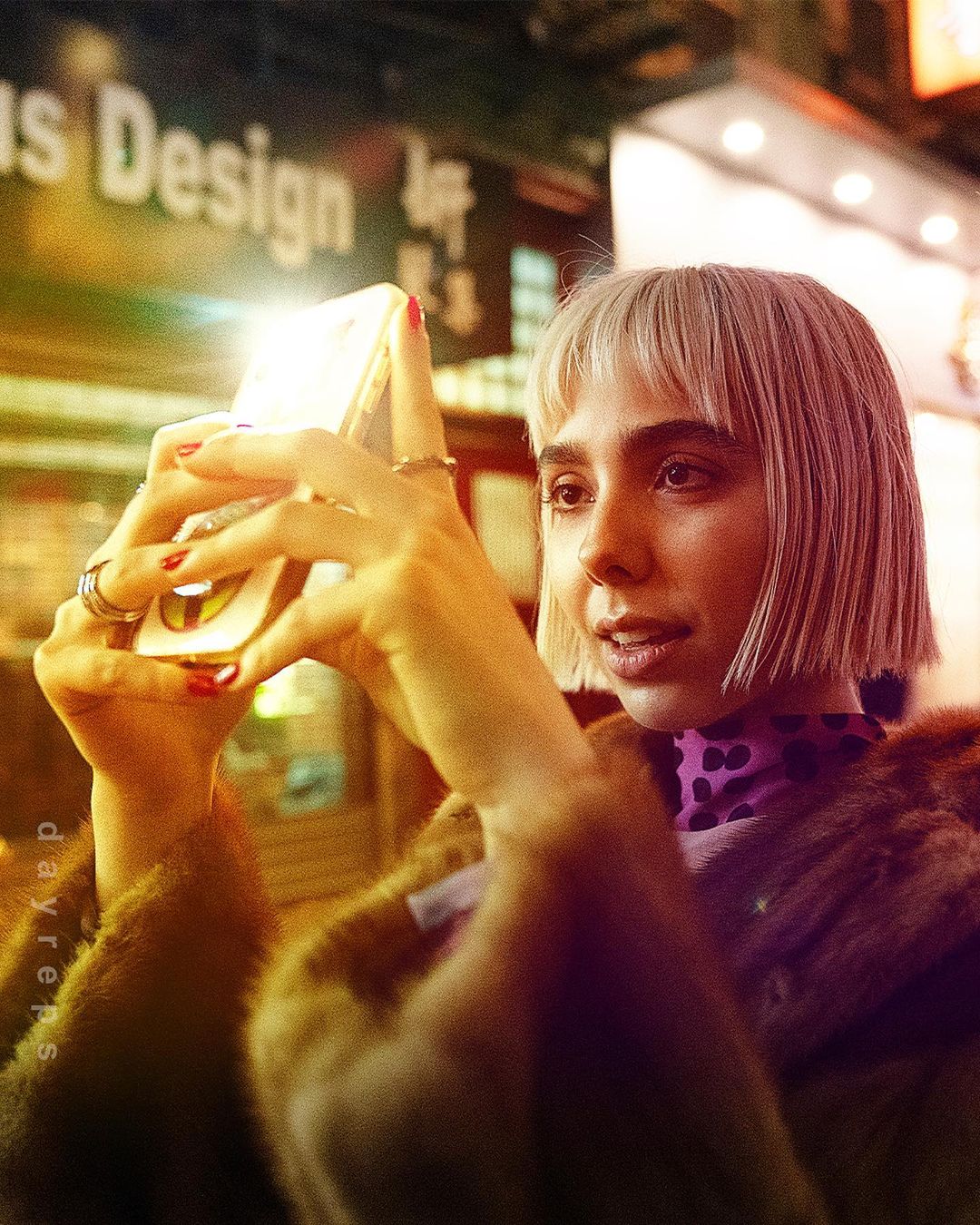 Blonde woman takes a photo with a smartphone in a nighttime urban setting, wearing a fur coat. Street style and modern technology.