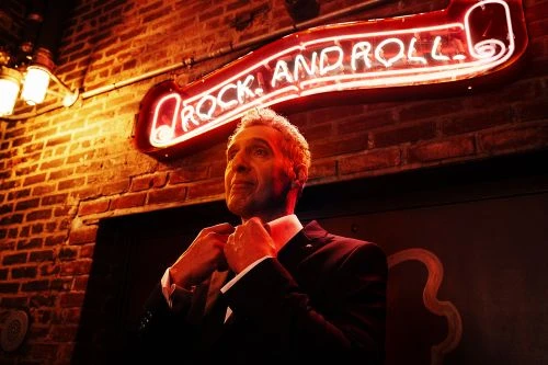 Adult male in a suit adjusts his bow tie in front of a red brick wall with a bright red neon sign reading "Rock and Roll."