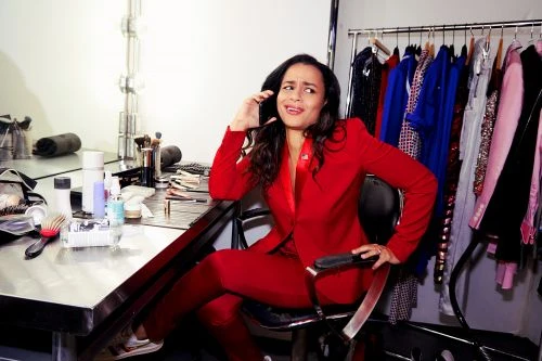 Adult female in a red suit is sitting at a makeup table using a cell phone, with a slight grimace, inside a dressing room.