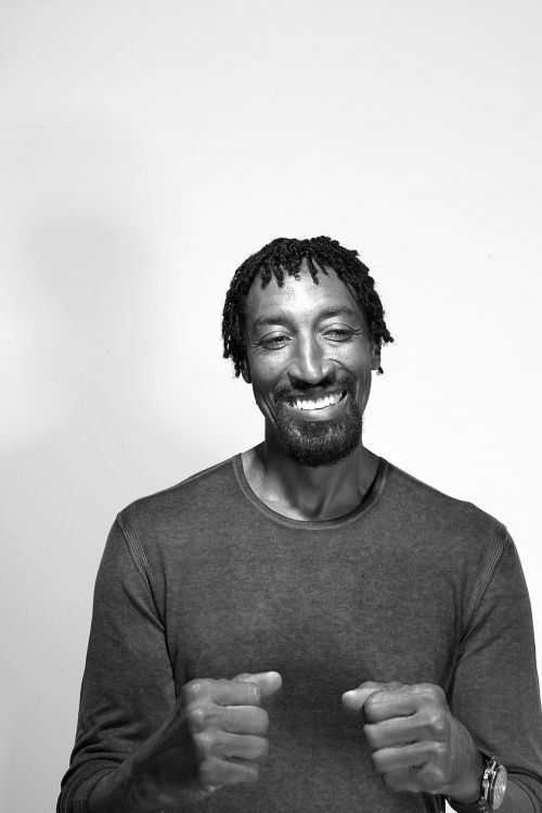 Black and white portrait of a smiling adult male with locs wearing a long-sleeved shirt against a plain white background in a photo studio.